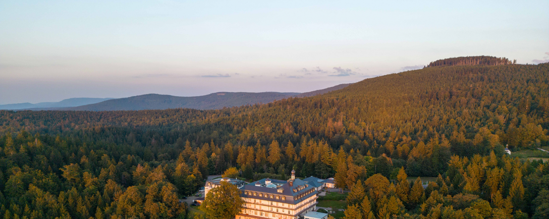 Luftaufnahme der Max Grundig Klinik, umgeben von dichtem Wald in hügeliger Landschaft bei warmem Abendlicht